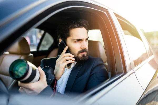 A person in a suit sits in a car, holding a camera and talking on a phone.