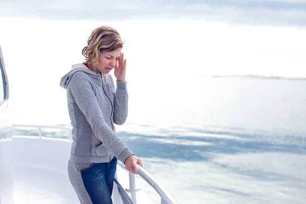 A person in a gray hoodie stands on a boat, looking at the water while resting one hand on their forehead.