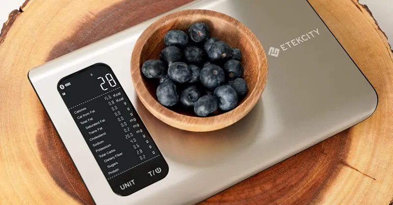 A digital kitchen scale displaying "28" grams with a small wooden bowl of blueberries on it, placed on a wooden surface.