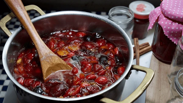 A saucepan filled with simmering plums and sugar, being stirred with a wooden spoon, next to empty jars and a covered jar on a wooden surface.