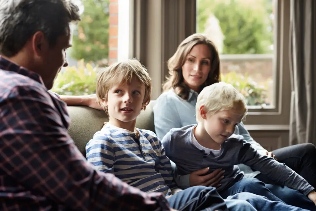 A family of four sits together on a couch in a sunlit room. Two children sit between a man and a woman, who appear to be engaged in conversation.