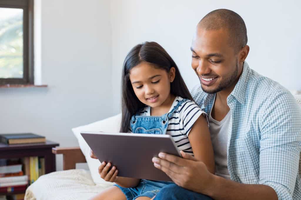 A man and a young girl sit together on a couch, smiling while looking at a tablet.
