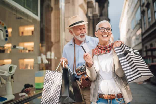An older couple happily holds shopping bags and a credit card while standing outside a store.