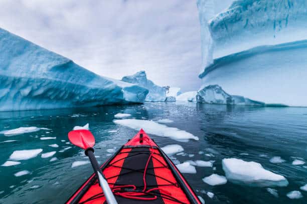 A red kayak navigates through icy waters surrounded by large blue icebergs under a cloudy sky.