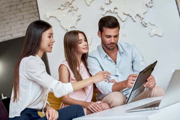 Three people collaborate while looking at a clipboard and laptop in an office with a world map on the wall.