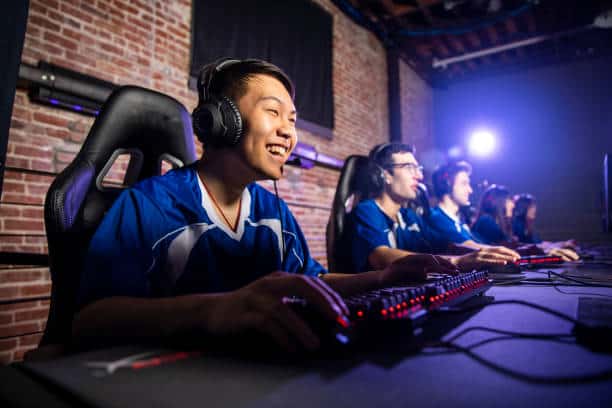 A group of people wearing headsets sit at computers in a dimly lit room, focused on gaming.
