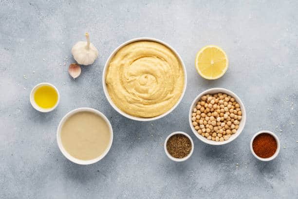 Bowls of hummus ingredients: chickpeas, tahini, lemon, olive oil, cumin, paprika, and garlic on a gray surface.