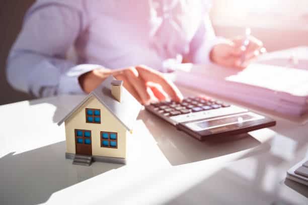 A person uses a calculator at a desk with a small model house in the foreground.