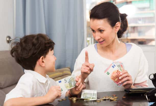 A woman and a child sit at a table with euro banknotes and coins. The woman gestures while holding money, and the child looks at her, smiling.