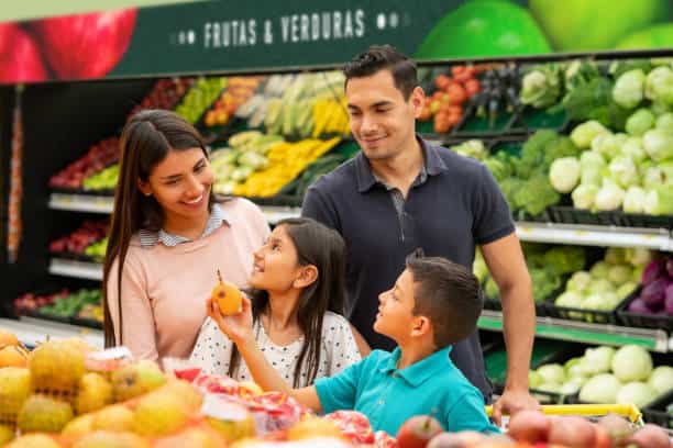 Family shopping for fruits and vegetables in a grocery store, with two children and their parents smiling and holding produce.