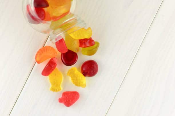 A jar spills colorful fruit-shaped gummy candies onto a white wooden surface.