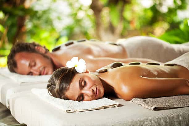 A man and a woman relaxing with hot stone therapy on their backs, lying on massage tables in a tranquil, leafy setting.