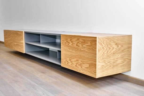 A minimalist wooden wall-mounted TV stand with open shelves and closed compartments, placed against a white wall on a wooden floor.