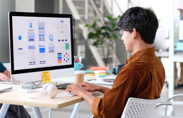 A person works on app design at a computer in a modern office setting with various design elements displayed on the screen.