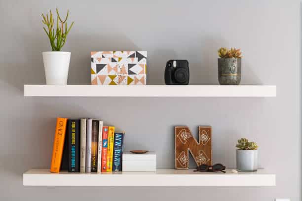 Two white shelves mounted on a wall; top shelf holds a potted plant, camera, and decor box; bottom shelf holds books, decorative letter 'M', sunglasses, and a small plant.
