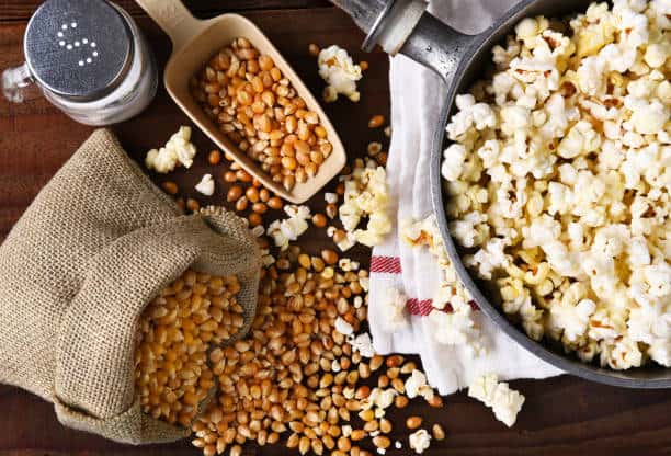 A bowl of popcorn beside spilled corn kernels and a burlap sack, with a salt shaker on a wooden table.