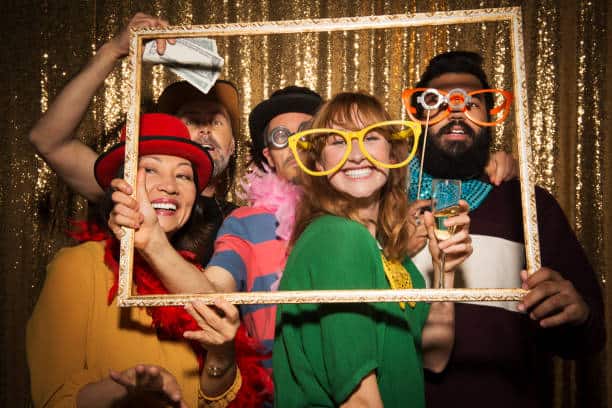 Five people wearing colorful accessories pose with a picture frame, standing in front of a gold sequin backdrop.