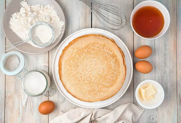 A stack of crepes on a plate surrounded by flour, sugar, eggs, butter, a whisk, and syrup on a wooden surface.