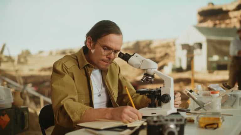 A person writing notes next to a microscope in an outdoor setting, with a camera and equipment nearby.