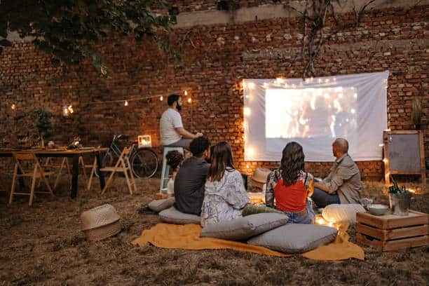 Four people sit outdoors watching a movie on a projector screen against a brick wall, surrounded by ambient string lights.