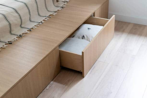 A wooden bed frame with an open drawer containing cushions on a light wood floor.