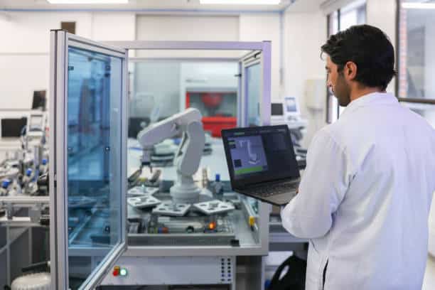 A man in a lab coat operates a laptop next to an industrial robot arm inside a laboratory.