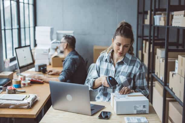A woman scans a package with a barcode scanner at a desk, while a man works on a computer in the background. The room is filled with boxes and a large window.