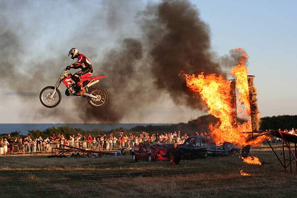 Motorcyclist performs a jump over a flaming obstacle as a crowd watches.