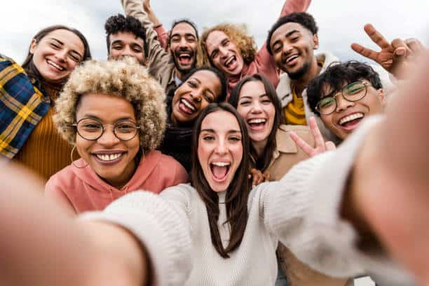 A diverse group of people standing close together outdoors, smiling and taking a selfie with outstretched arms.