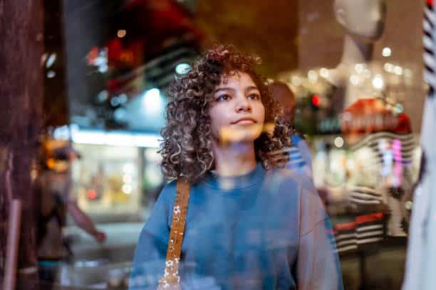 Young woman with curly hair looks through a shop window at night, wearing a blue sweatshirt, with reflections of city lights and mannequins visible on the glass.
