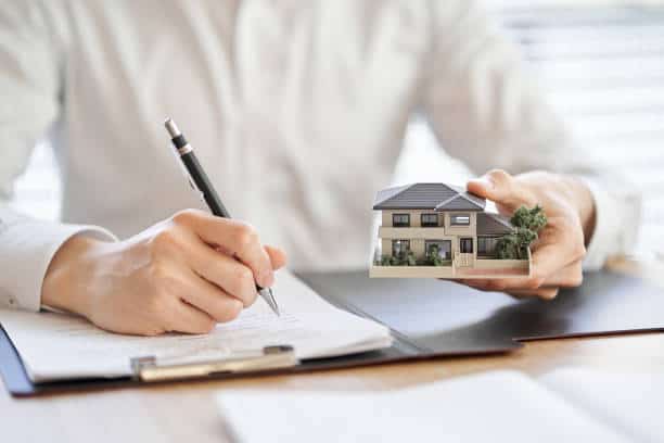 Person holding a house model in one hand and writing on a clipboard with the other hand.