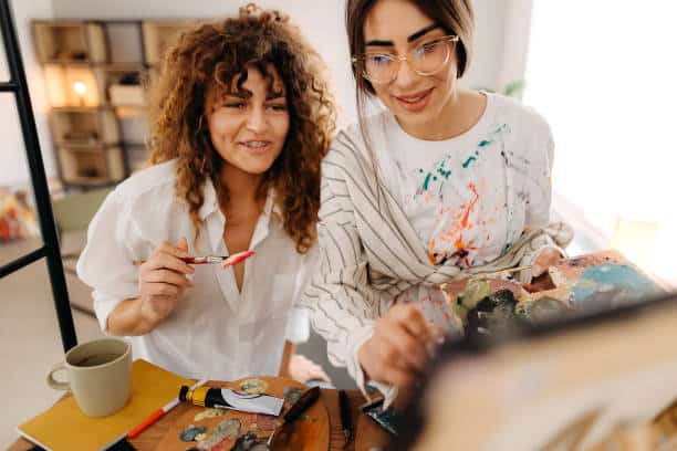 Two women painting together, surrounded by art supplies. One holds a palette, while the other holds a brush. They appear engaged and focused on their work.