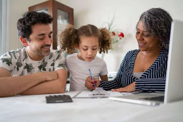 A child writes on paper while seated at a table with two adults. A laptop and a calculator are on the table. The adults observe and smile.