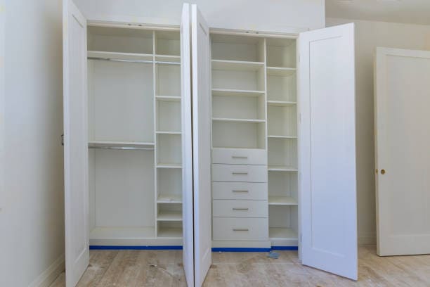Open white closet with multiple shelves and drawers in an empty room with wooden flooring.