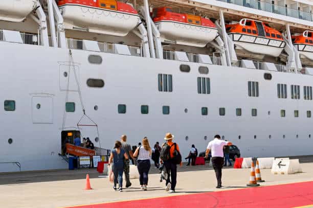 People walking toward the entrance of a docked cruise ship with visible lifeboats on the deck.