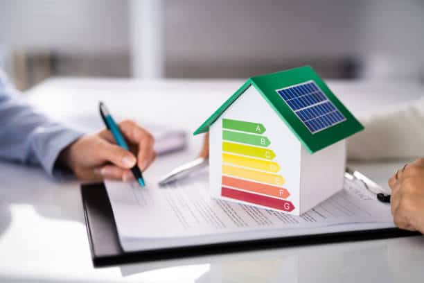 A miniature house with an energy efficiency rating label and a solar panel sits on a document. Two people are partially visible, one holding a pen.