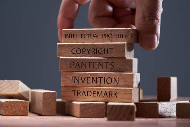 A hand stacks wooden blocks labeled "Intellectual Property," "Copyright," "Patents," "Invention," and "Trademark" on a table.