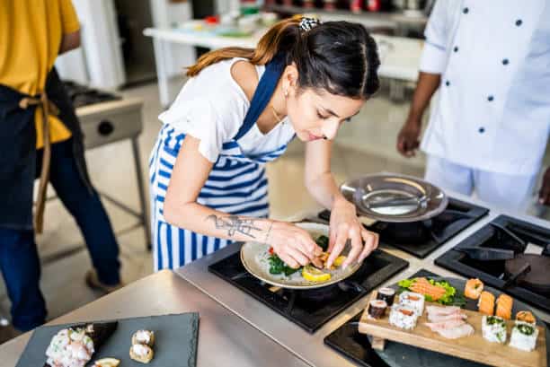A woman in a striped apron carefully plates food in a kitchen, while a chef in white stands nearby. Sushi dishes are prepared on the counter.
