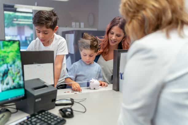 A woman and two children are at a bank counter. The children are placing coins into a piggy bank. A bank employee is visible from behind the counter.