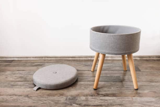 A gray, round pet bed with wooden legs stands on a wooden floor. A matching gray cushion lies beside it.