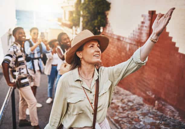 A woman in a hat gestures while leading a group of people on a sunny street. The group seems to be enjoying a guiding tour or walk.