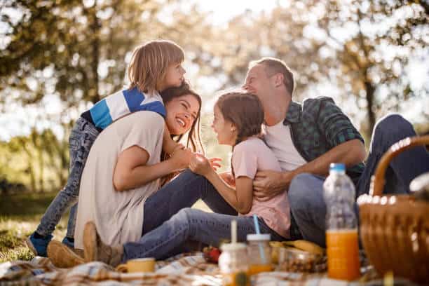 A family of four enjoys a picnic on a blanket in a sunny park, smiling and laughing together.