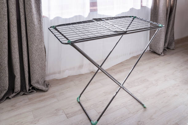 A metal clothes drying rack stands unfolded on a wooden floor near curtained windows.