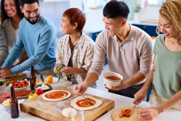 Group of people making pizzas together at a kitchen counter, with various toppings and ingredients laid out.