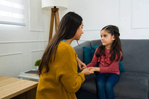 A woman in a yellow sweater is kneeling and holding the hands of a young girl in a pink shirt seated on a gray couch, in a room with white walls and a floor lamp.