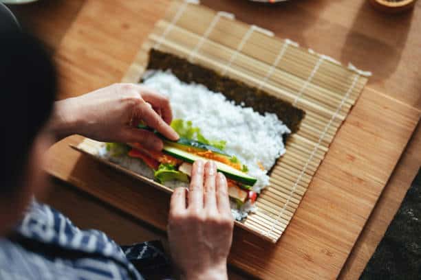 Hands rolling a sushi roll with rice, vegetables, and seaweed on a bamboo mat.