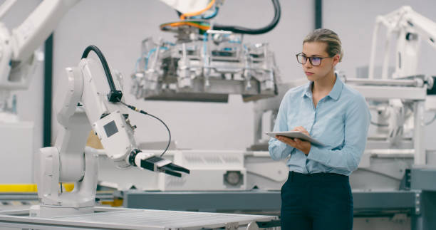 A person with glasses is standing beside a robotic arm, holding a tablet, in an industrial setting.
