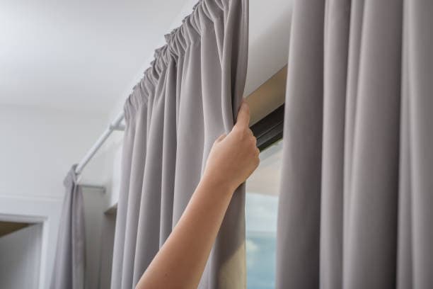 A hand pulling back a gray curtain in front of a window.