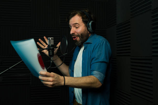 A man in a recording studio holds a script, wearing headphones and speaking into a microphone, surrounded by acoustic foam panels.