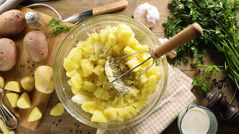Top-view of a bowl with mashed potatoes and a masher. Surrounding are whole and cut potatoes, garlic, salt, a knife, fresh herbs, and cream on a wooden table.